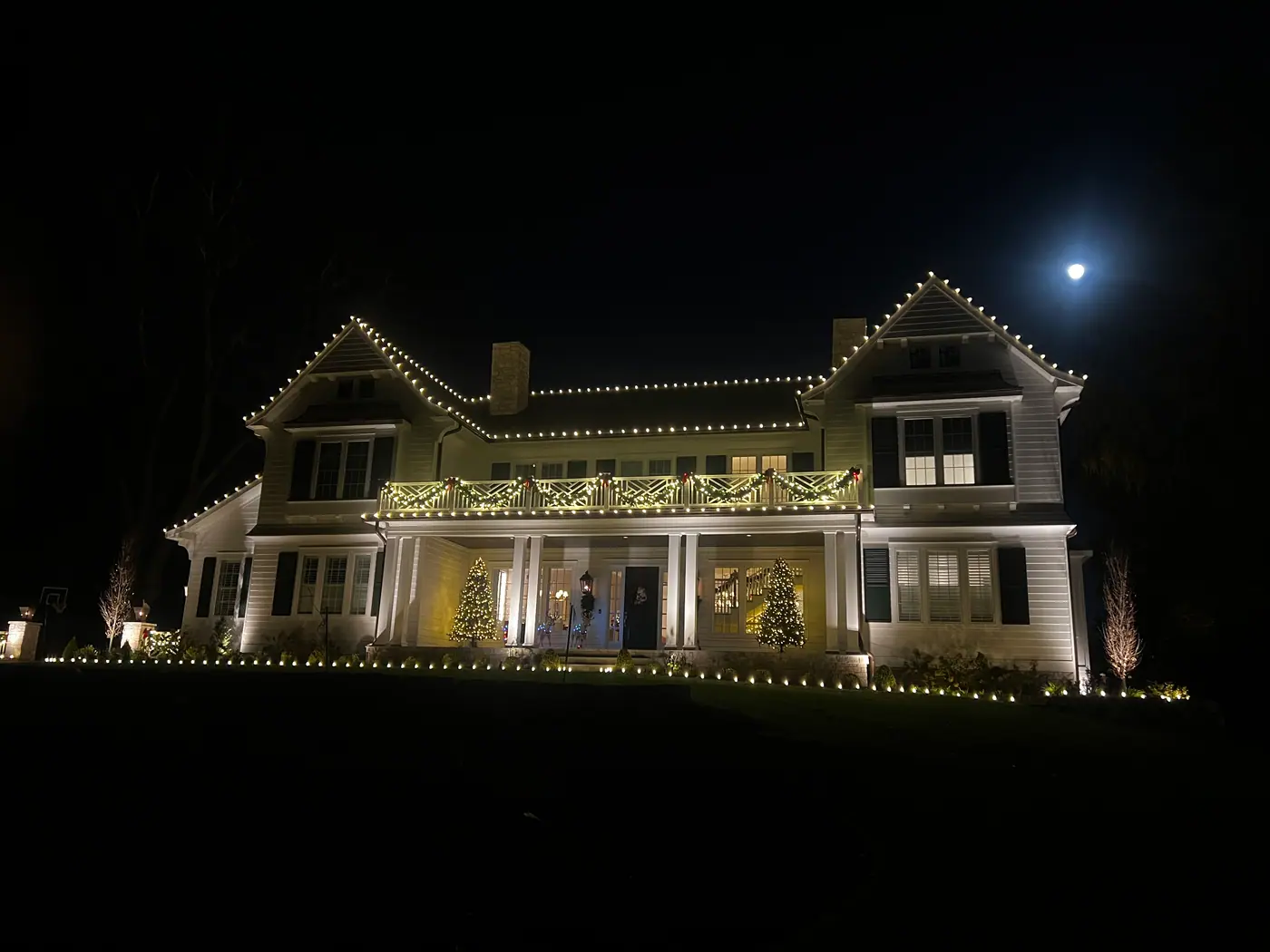 The same house in December, roofline traced in warm white with lit garland along the balcony and trees on the porch