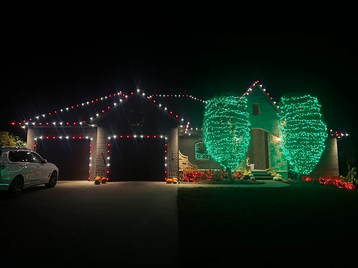 A home with red and white roofline lights and two large trees wrapped in brilliant green